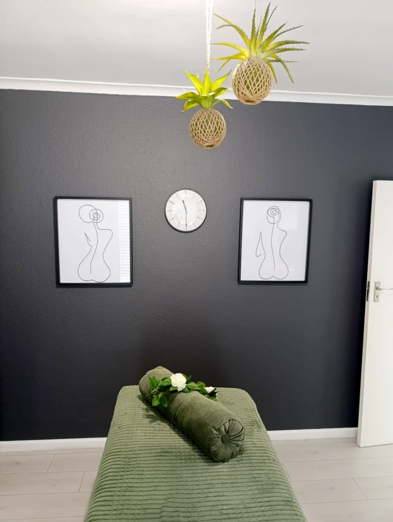 Modern treatment room with a dark feature wall, minimalist art, and a green massage bed decorated with white flowers.